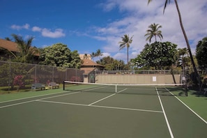 A tennis court with a net, surrounded by a fence, palm trees, and a building in the background. - Kihei Bay Surf 227 by Maui Rental Group (Kihei)