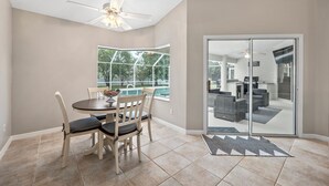 A dining area with a table and chairs, a ceiling fan, a large window, and a sliding glass door leading to a living area. - Summer Daze by Distinctive Beach Rentals (Palm Coast)