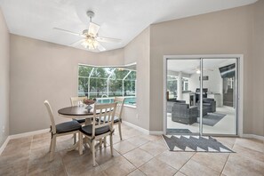 A dining area with a table and chairs, a ceiling fan, a large window, and a sliding glass door leading to a living area. - Summer Daze by Distinctive Beach Rentals (Palm Coast)