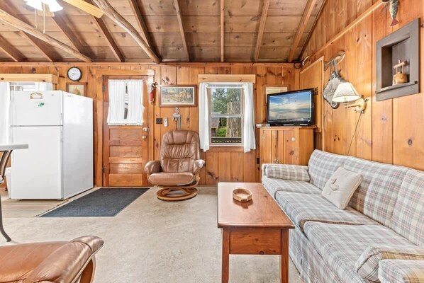 L’intérieur chaleureux d’un chalet, avec un revêtement mural en bois, un canapé, un fauteuil, un téléviseur et un réfrigérateur.