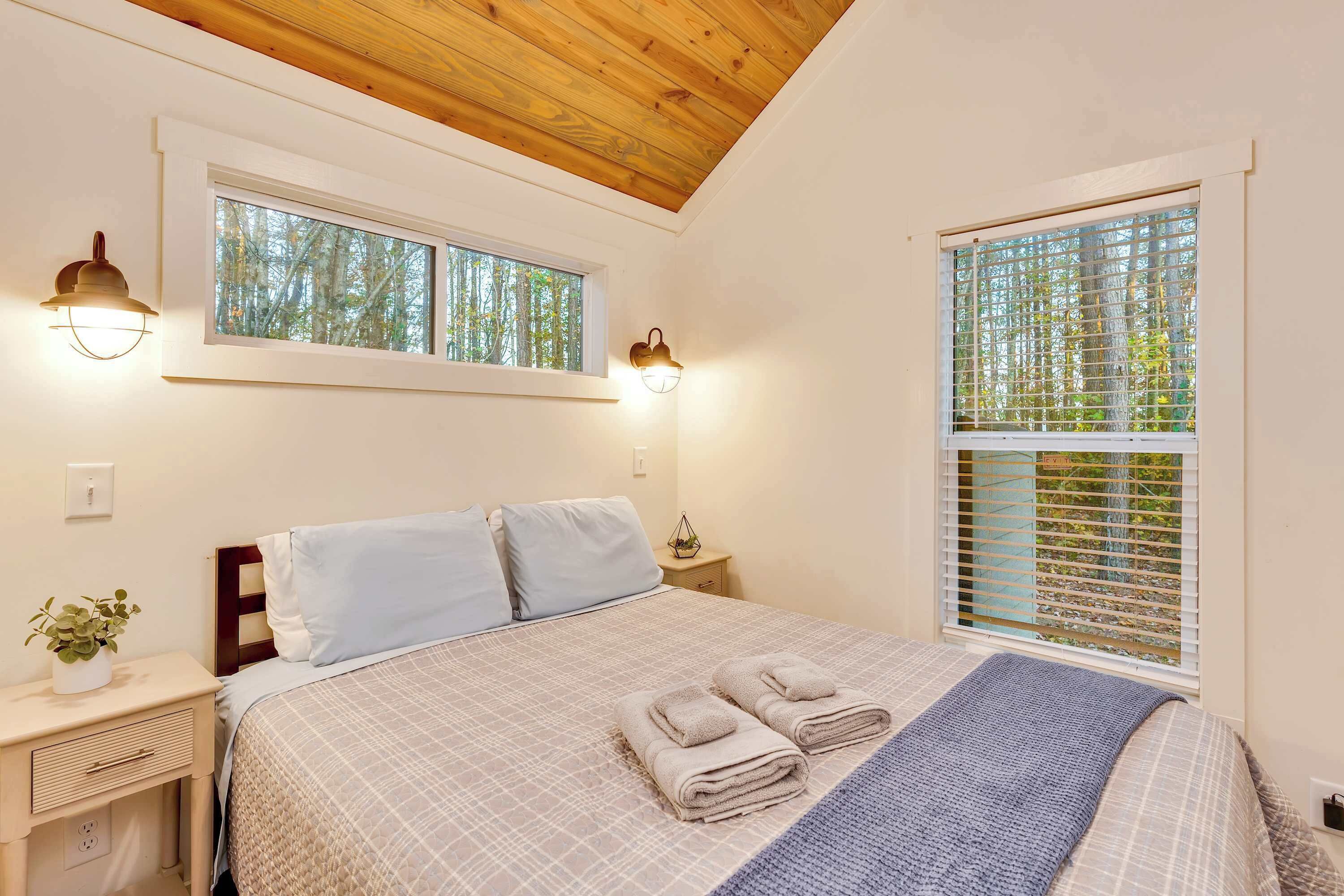 A neatly made bed with a checkered bedspread and two folded towels, a bedside table with a lamp, and a window with blinds.