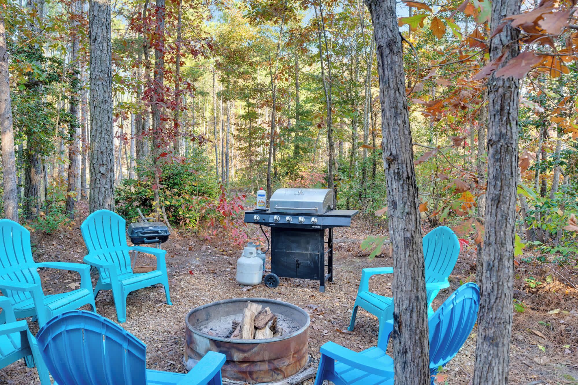 A forest setting with a wood-burning stove, a grill, and blue chairs arranged around a fire pit.