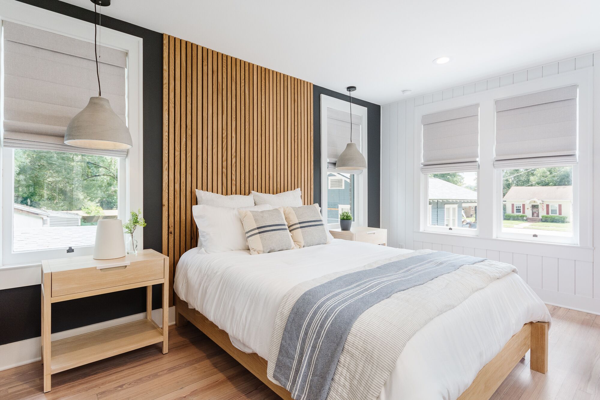 A modern bedroom with a wooden headboard, a bedside table with a lamp, and large windows with blinds.