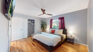 Een slaapkamer met een bed, een plafondventilator, een raam met gordijnen, een nachtkastje met een lamp en een garderobe.