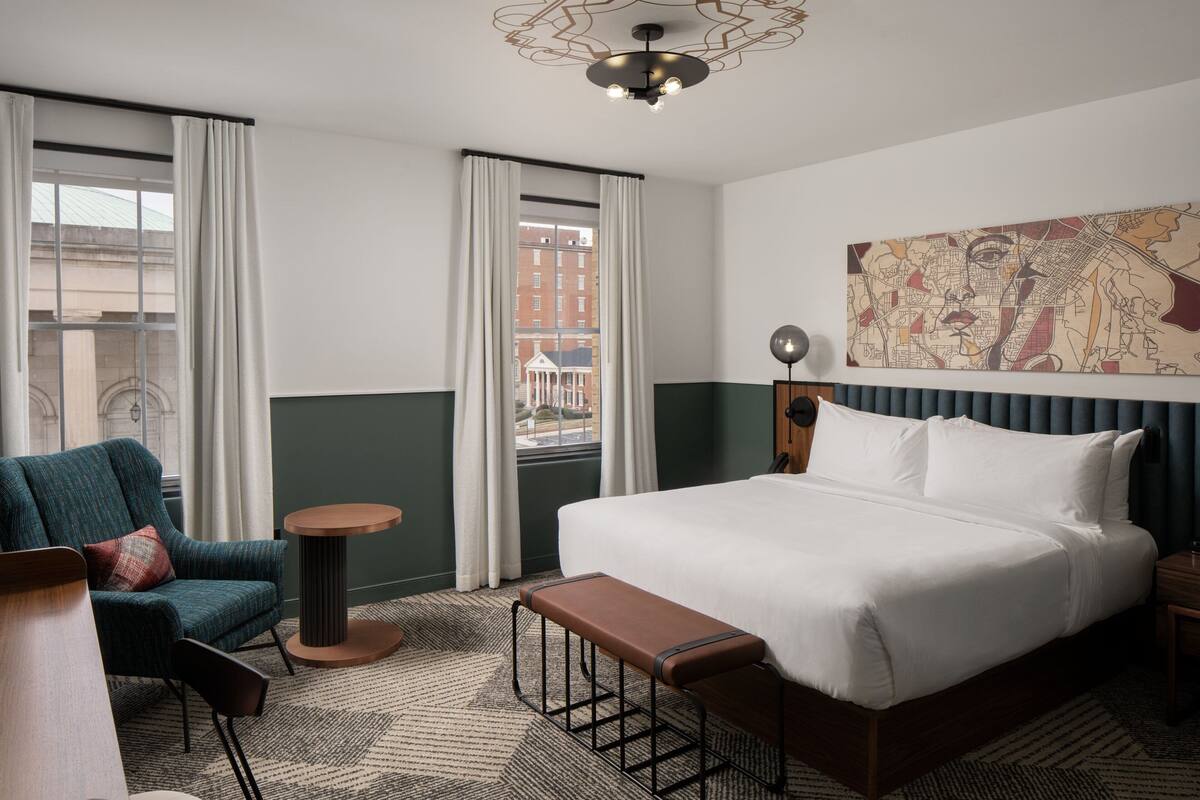 A hotel room with a large bed, a bench, a chair, a round table, and a cityscape view through the window.