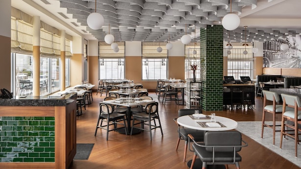Un restaurant moderne avec de hauts plafonds, des murs en briques apparentes et différents types de sièges, notamment des tables rondes et des banquettes.