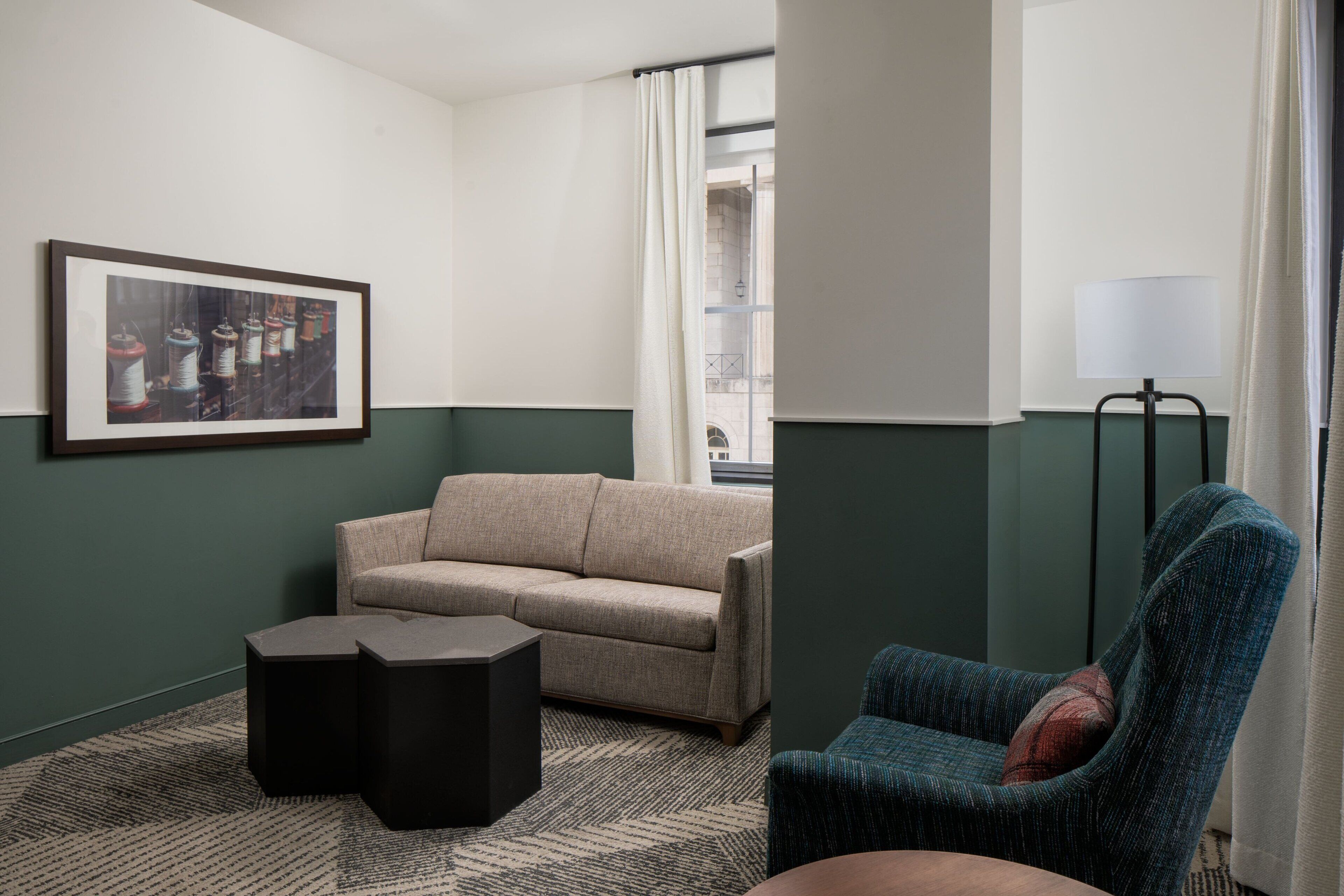 A room with a beige sofa, a black ottoman, a framed textile artwork, a blue armchair, and a floor lamp.