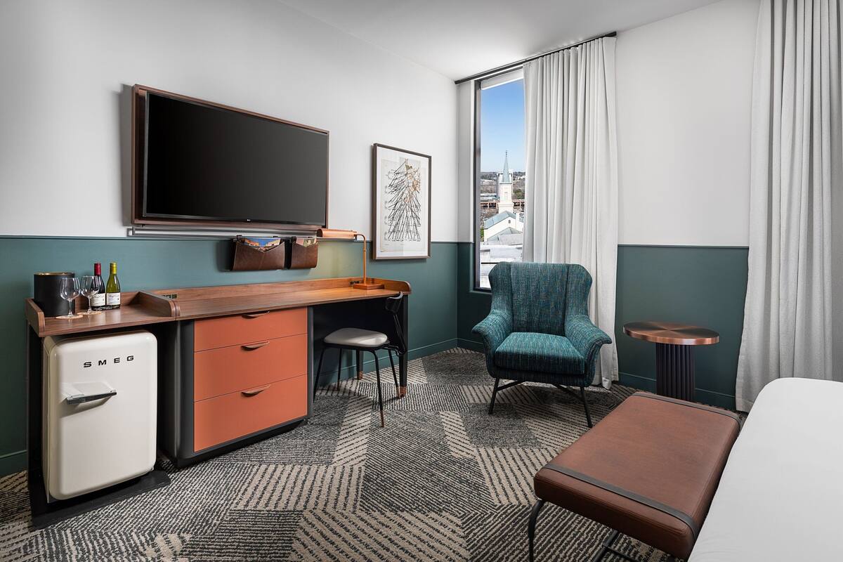 A hotel room with a minibar, a desk, a chair, a bed, and a window with curtains.