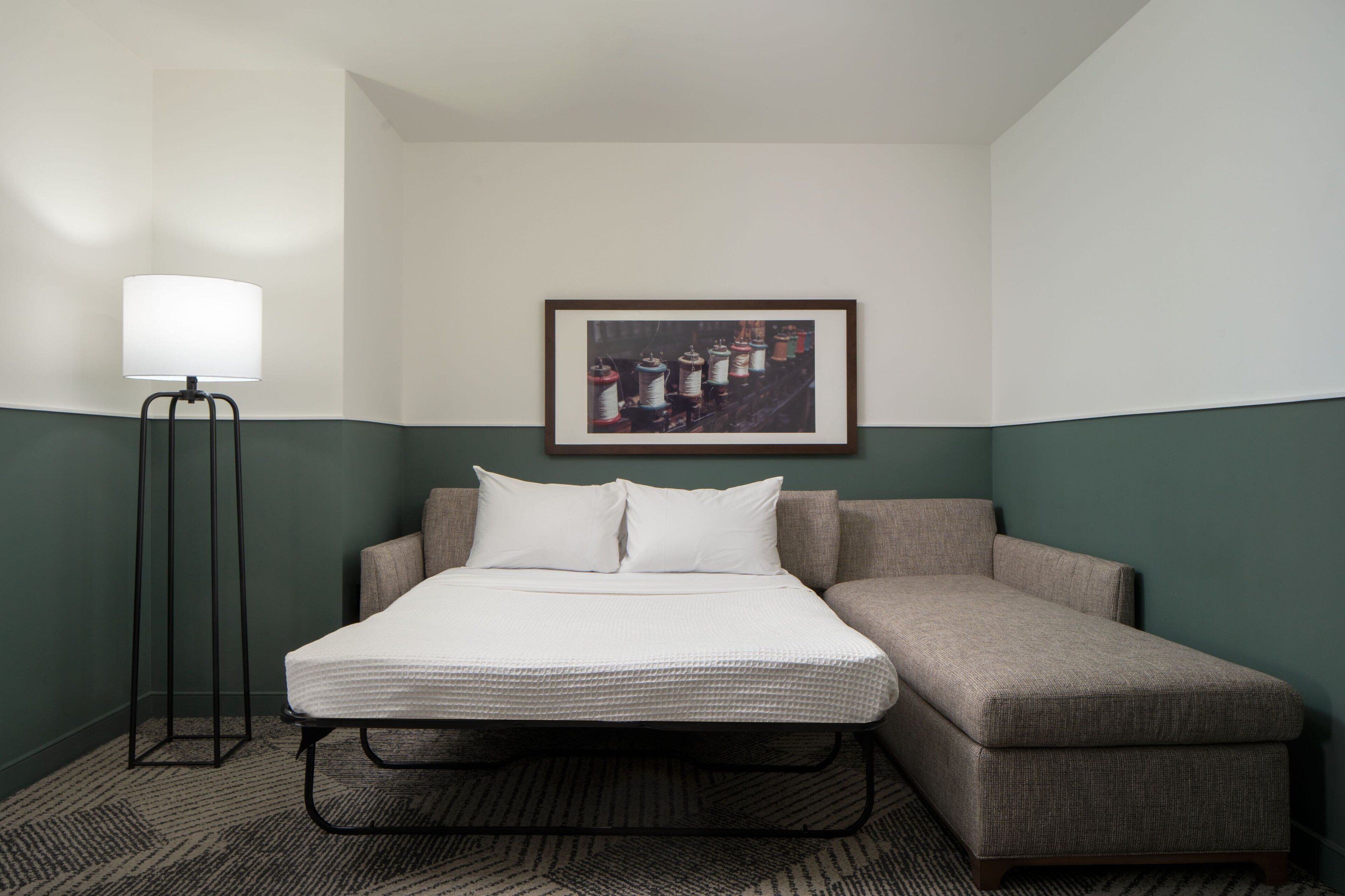 A hotel room with a bed, a floor lamp, and a framed picture on the wall.