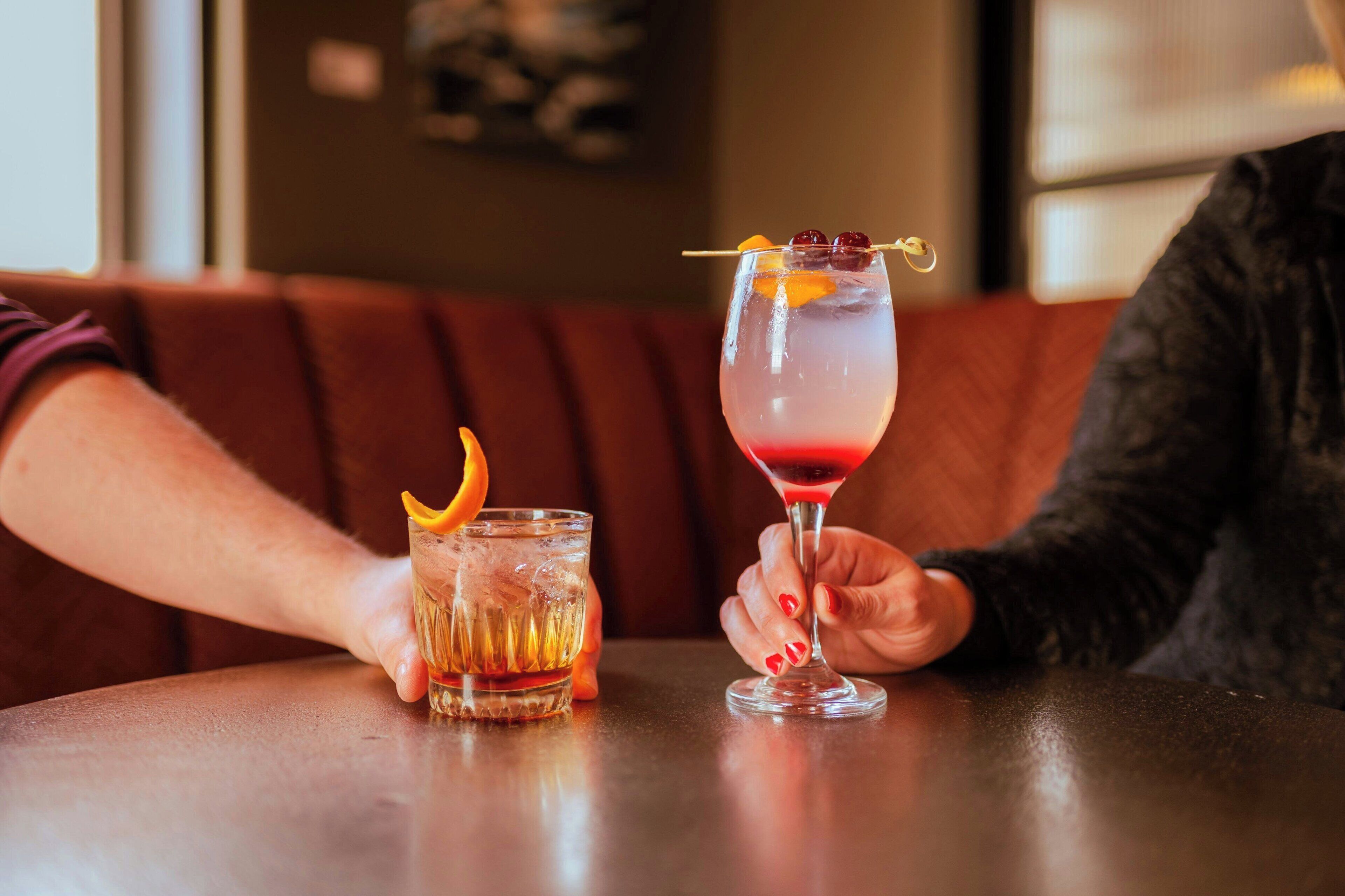Two people seated at a table with two drinks, one in a short glass and one in a martini glass, with a red drink and garnishes.