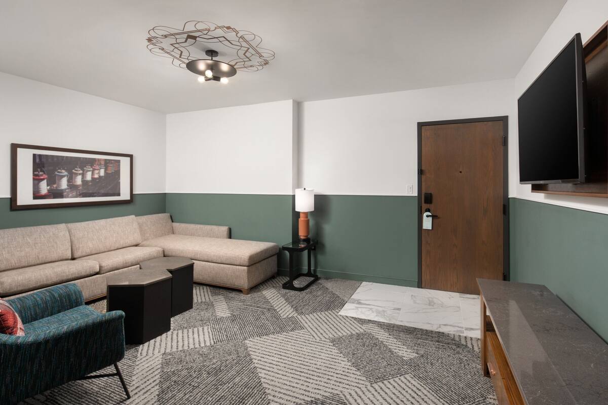 A modern living room with a sectional sofa, a small side table, a patterned rug, a wall-mounted TV, and a door with a wooden frame.