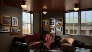 Un salon moderne avec un canapé d’angle, des fauteuils et une petite table. Les murs sont ornés de tableaux encadrés et de grandes fenêtres offrent une vue sur la ville.