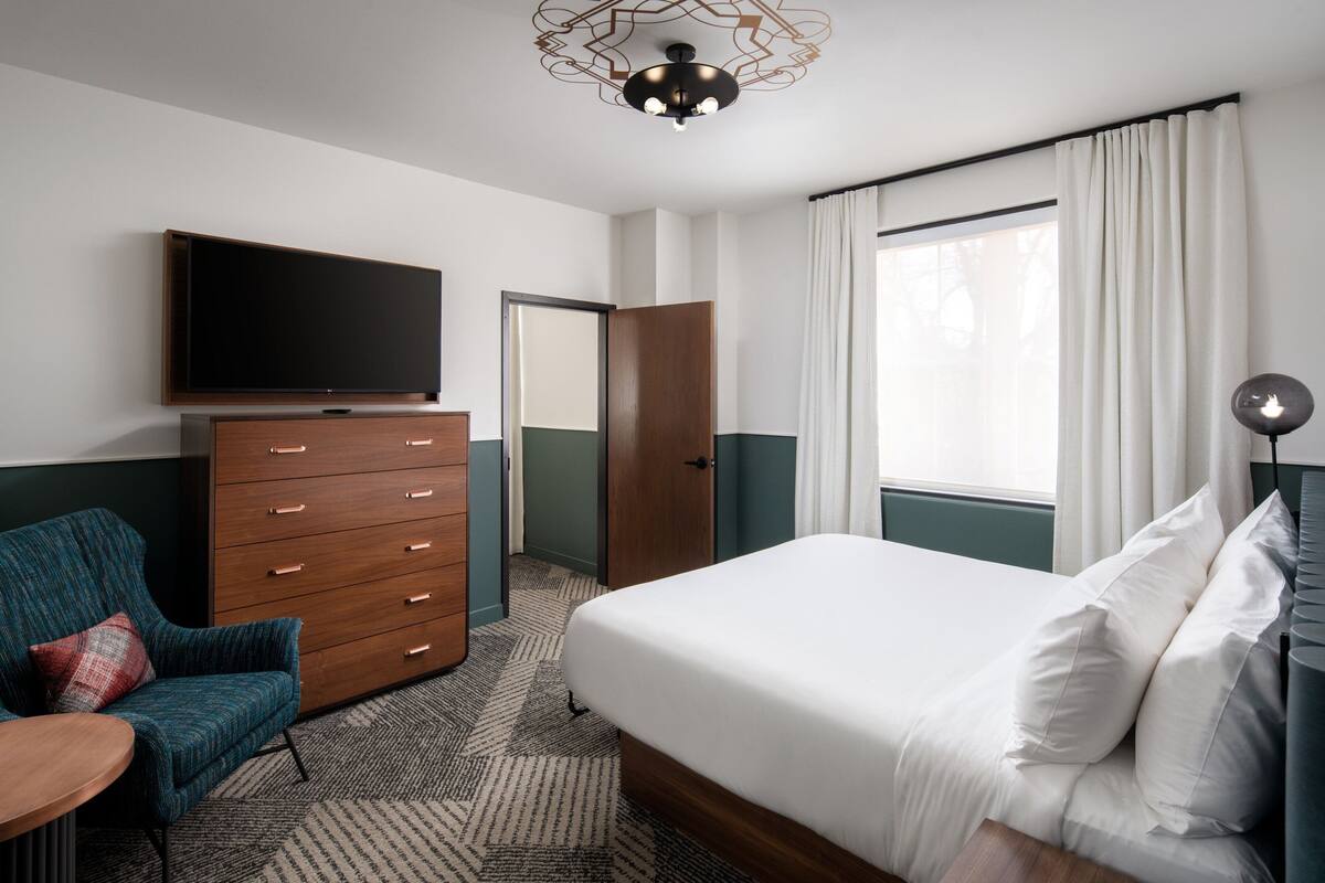 A hotel room with a bed, a TV, a dresser, a chair, and a window with curtains.