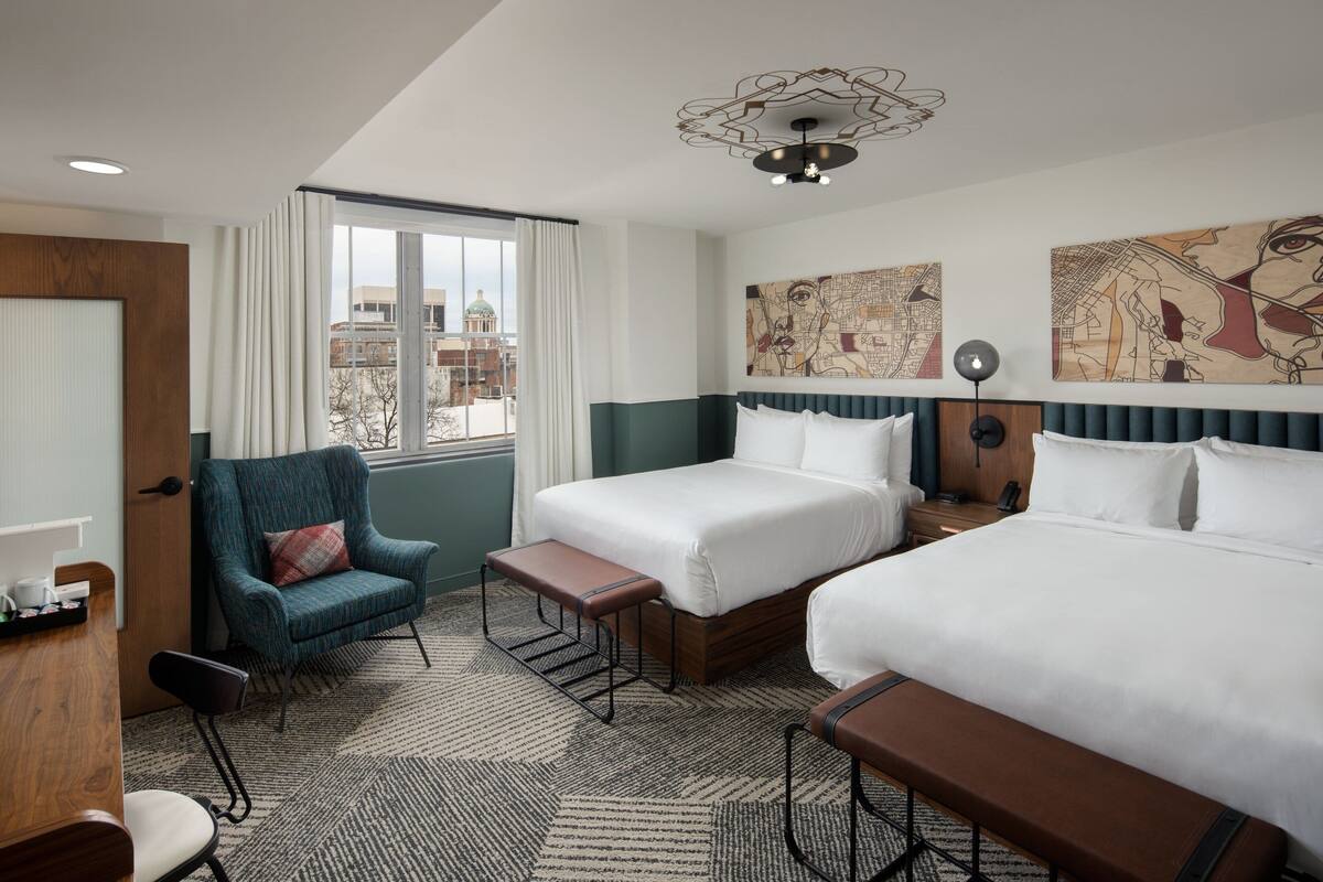 A hotel room with two beds, a chair, a desk, and a view of the city.