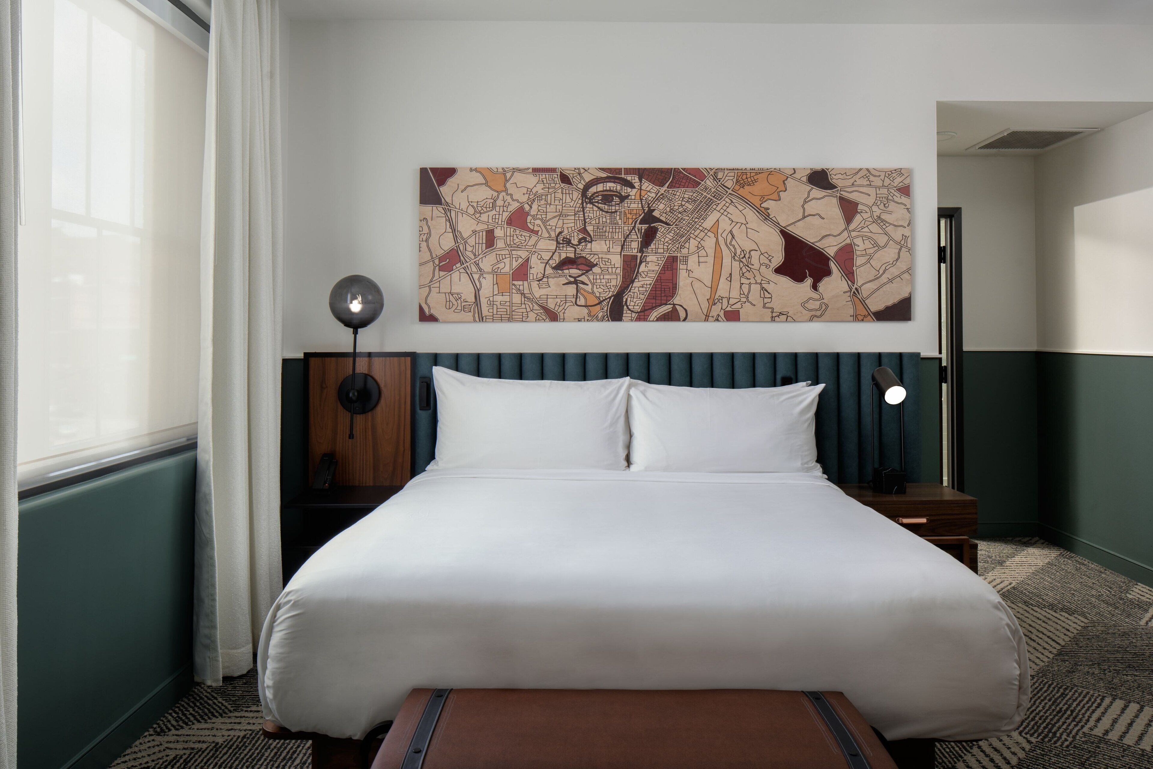 Quarto de hotel com uma cama grande, um quadro com um mapa na parede, uma mesa de cabeceira com abajur e vista para a cidade pela janela.
