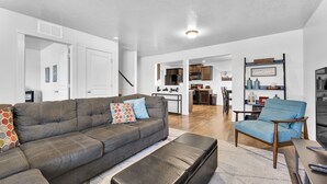 A modern living room with a grey sofa, a blue armchair, and a black ottoman. There is a kitchen in the background with wooden cabinets and a dining area. - Sweet as a Peach Family Home (Meridian)