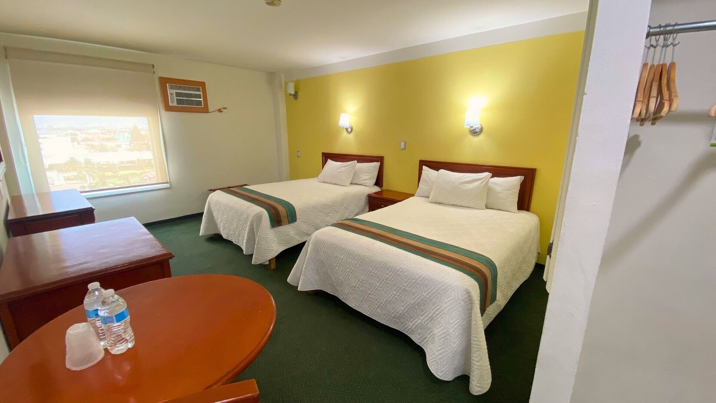 Habitación de hotel con dos camas, una mesa de madera con botellas de agua, una ventana y un aire acondicionado instalado en la pared.