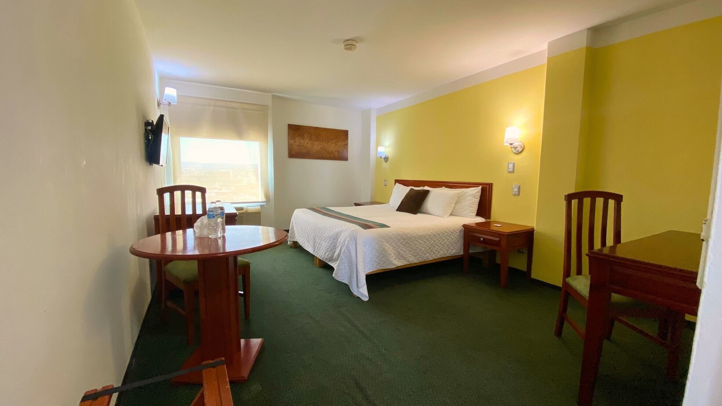 Habitación de hotel con cama, una mesita, dos sillas, ventana con persianas y un cuadro en la pared.