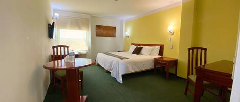 Habitación de hotel con cama, una mesita, dos sillas, ventana con persianas y un cuadro en la pared.
