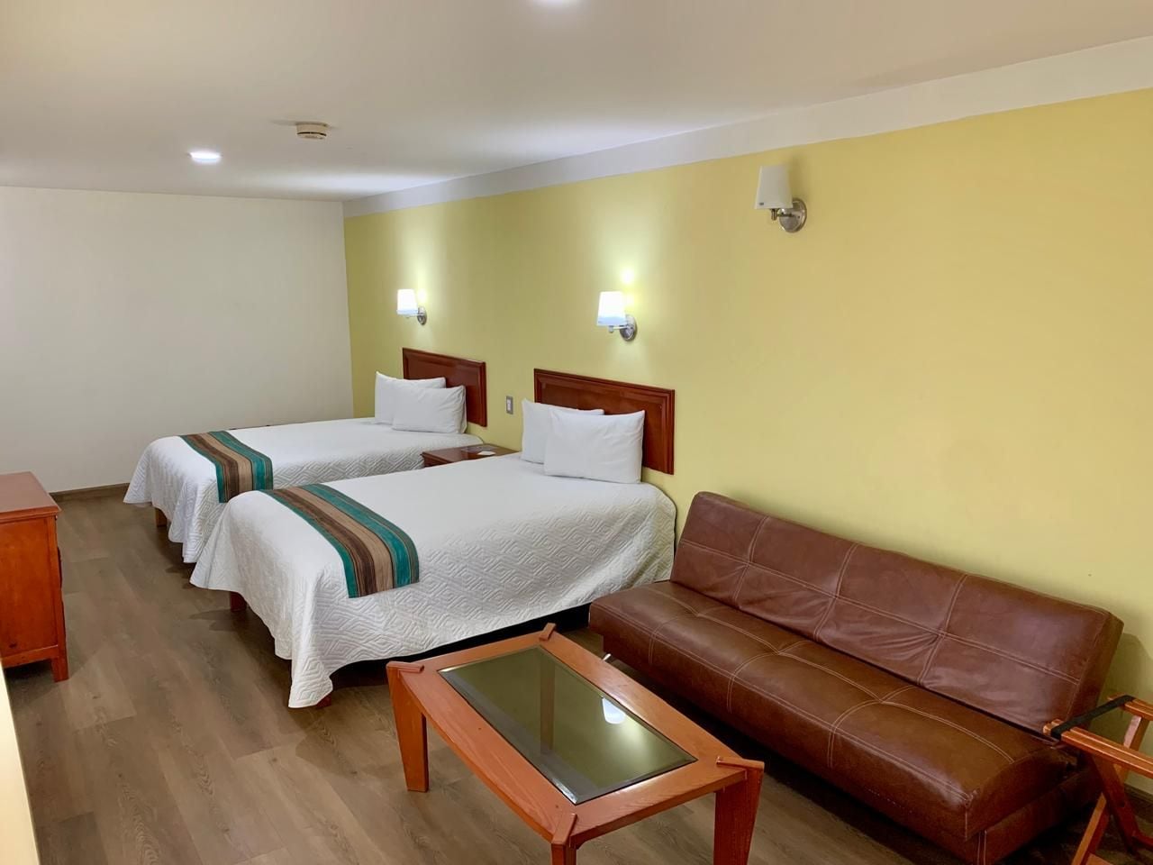 A hotel room with two single beds, a brown leather sofa, a wooden coffee table, and a small wooden side table.