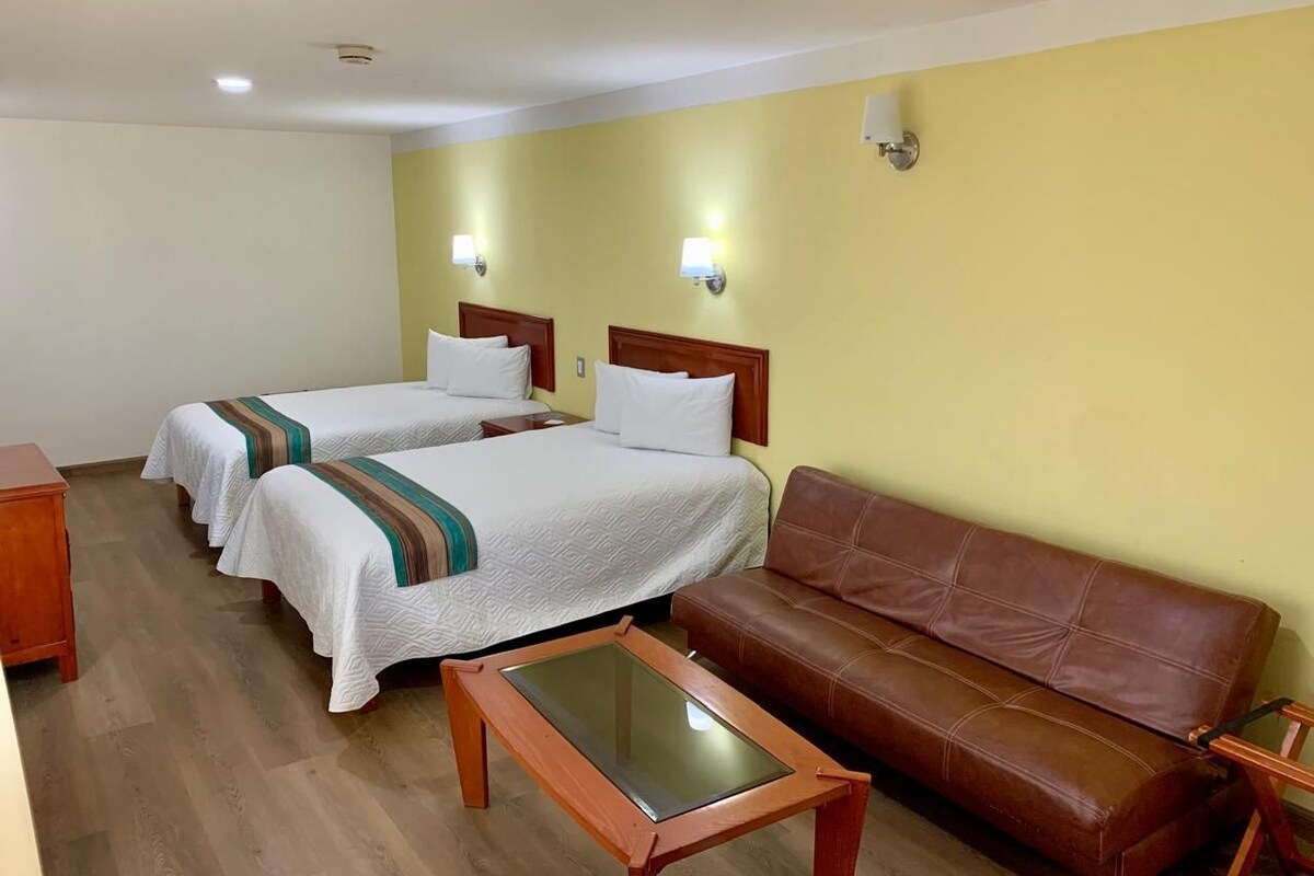 Una camera d'albergo con due letti singoli, un divano in pelle marrone, un tavolino in legno e un piccolo comodino in legno.