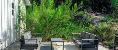 An outdoor patio with two grey sofas and a small table, surrounded by dense green foliage.
