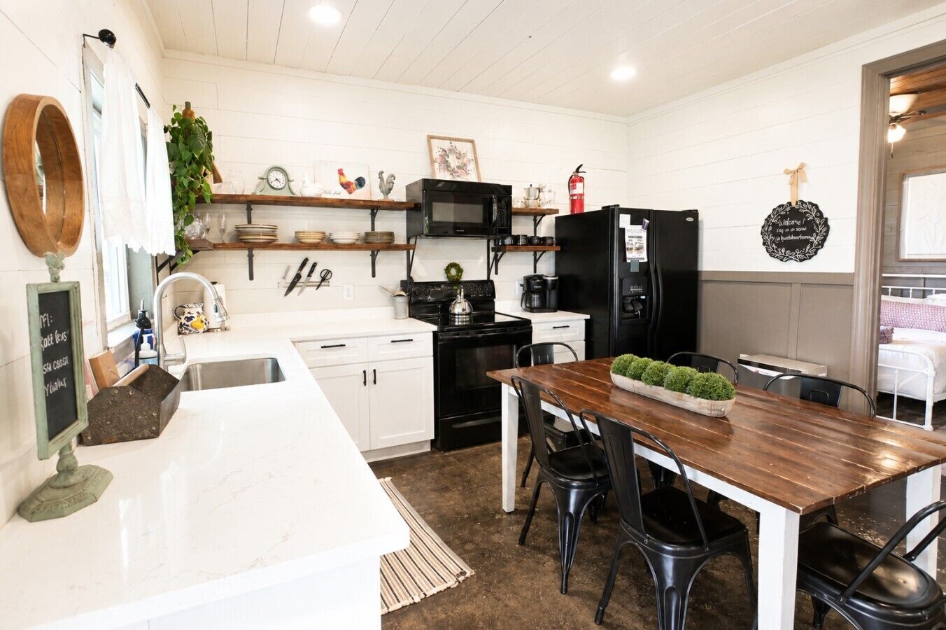 Une cuisine moderne avec des meubles blancs, une table à manger en bois, des chaises noires, un réfrigérateur noir et un four à micro-ondes.