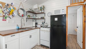 A compact kitchen with white cabinets, a black refrigerator, a stove, and a microwave. There are shelves with plants and a corkboard with various items pinned to it. - Summer Escape with Swim Hole and Golf Cart Rental (Spring Branch)
