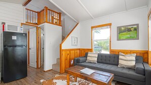 A cozy living room with a grey sofa, wooden coffee table, and a cowhide rug. A black refrigerator is on the left, and a wooden staircase leads to an upper level. - Summer Escape with Swim Hole and Golf Cart Rental (Spring Branch)