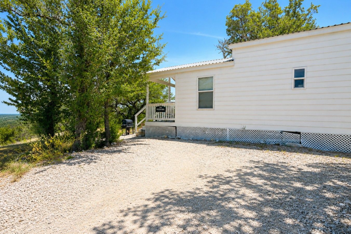 A single-story mobile home with a gravel driveway, a fenced patio area, and a view of the surrounding landscape.