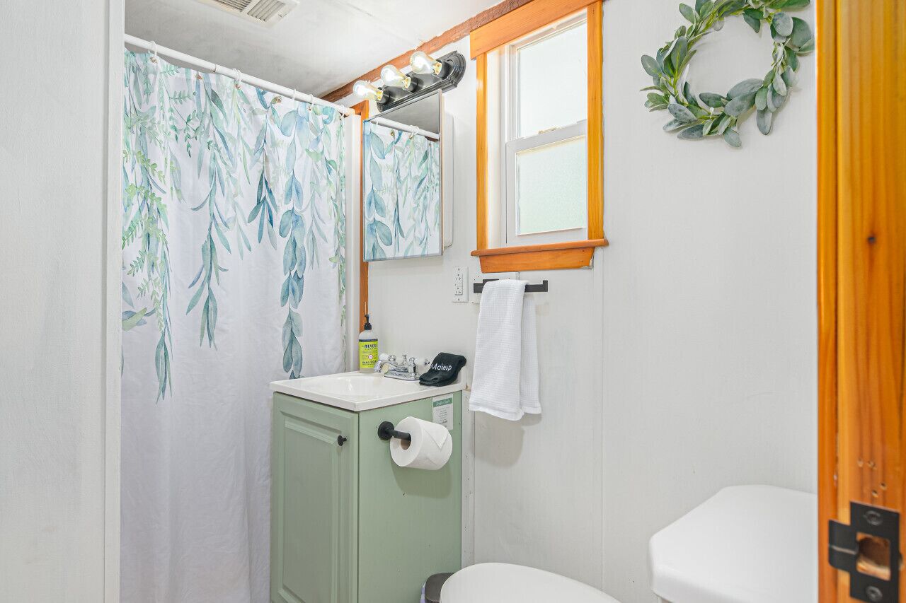 A bathroom with a green cabinet, a white toilet, a shower curtain with a leaf pattern, a mirror, and a window with a wreath.