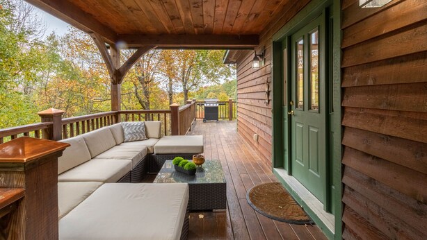 Eine Holzterrasse mit einer Couch und einem Tisch, eine grüne Tür und Blick auf Bäume.