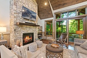 A cozy living room with a stone fireplace, comfortable seating, and large windows offering a view of the outdoors.