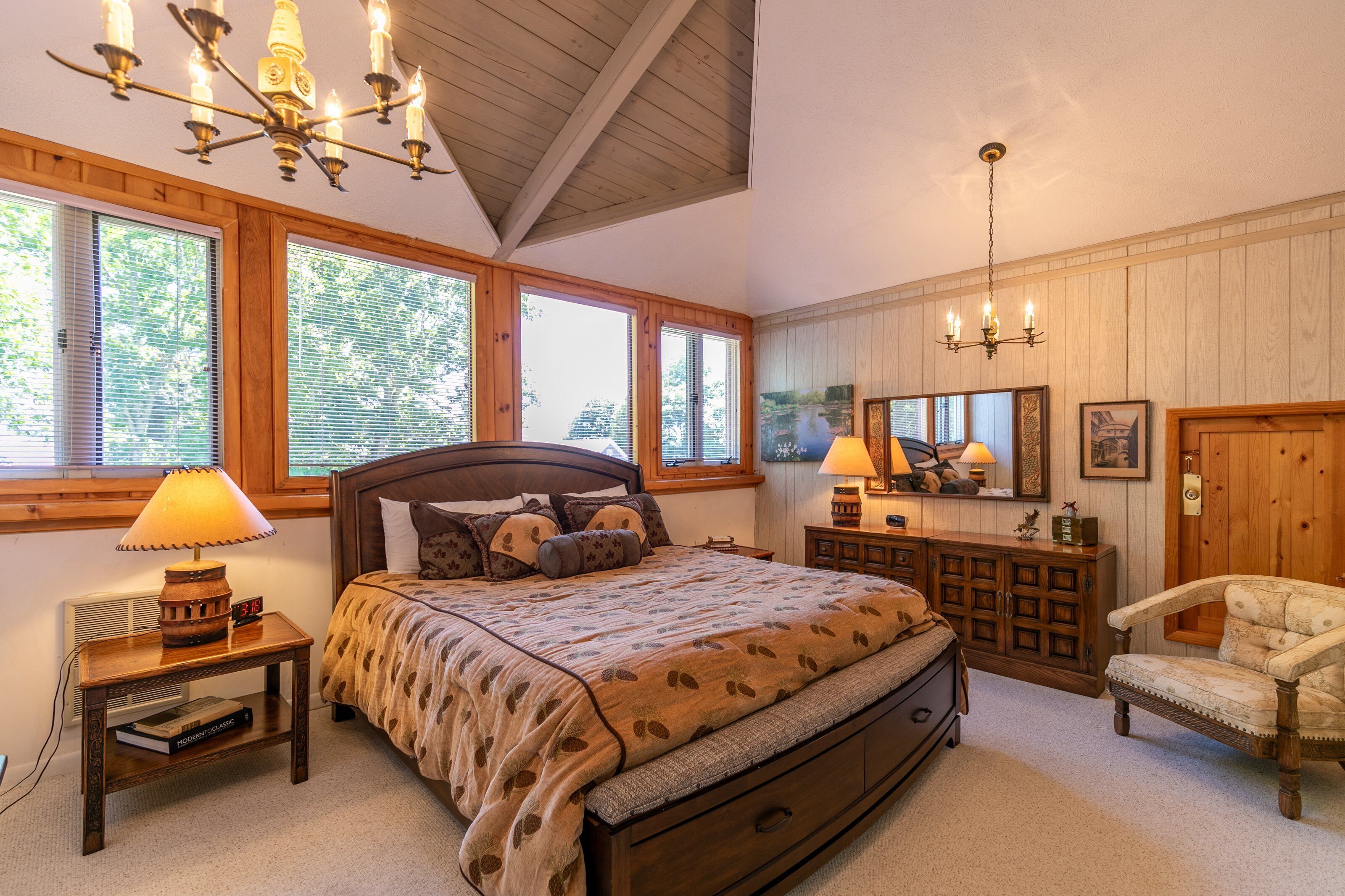 A bedroom with a large bed, wooden headboard, and a chandelier. There are two bedside tables with lamps and a dresser with a mirror.