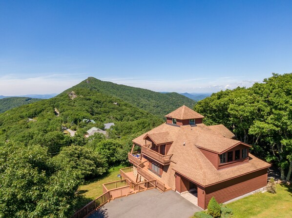 A two-story wooden cabin with a balcony, surrounded by lush green forest and a mountain in the background.