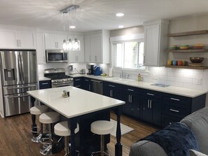 A modern kitchen with white cabinets, stainless steel appliances, a central island with stools, and a window providing natural light. - Elvis was here in 1956! (Biloxi)