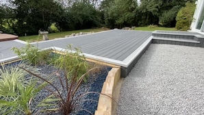 Eine Terrasse mit Holzboden, umgeben von einem Kiesstreifen und einem Holzpflanzkasten mit Pflanzen.