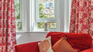A red sofa with decorative pillows, a white window with a view of trees, and red and white floral curtains. - Home-from-home; 10 mins from city centre; Free on-street parking; Well-equipped (Edinburgh)