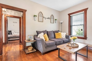 A living room with a grey sofa, wooden flooring, a small table with a vase of flowers, and a window with blinds.