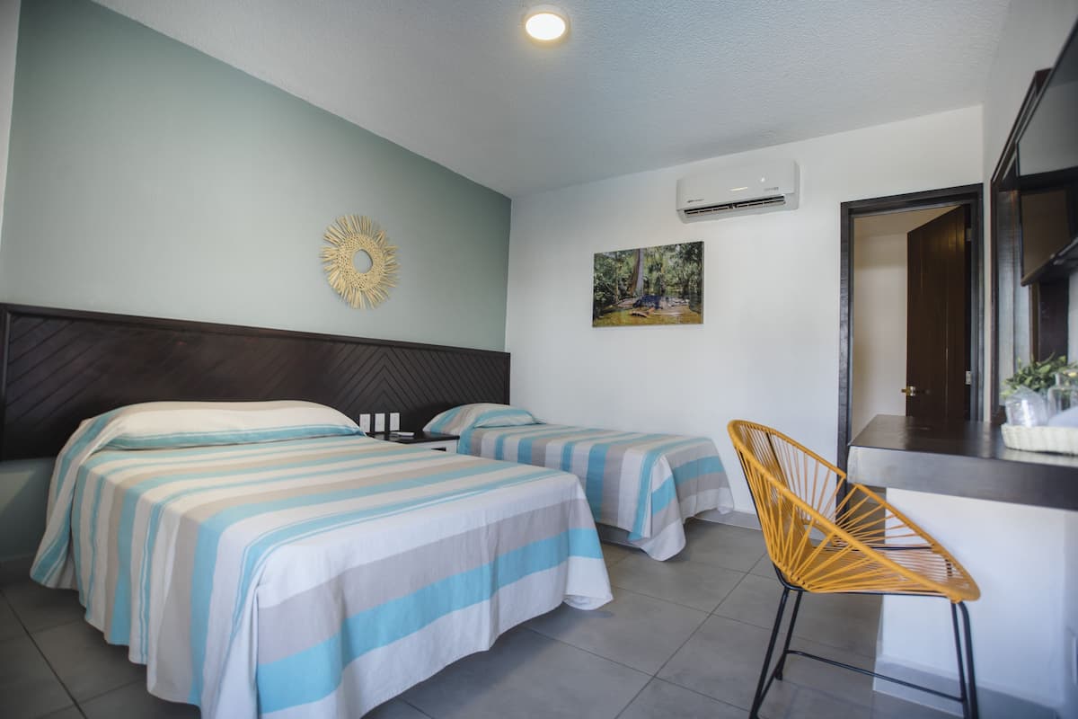Une chambre d’hôtel avec deux lits, une tête de lit en bois, un climatiseur fixé au mur, un miroir décoratif en forme de soleil et une petite chaise en bois.
