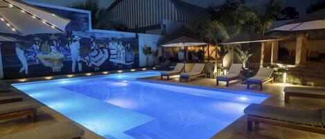 Una piscina con sillas de descanso y sombrillas, rodeada por un mural y palmeras.