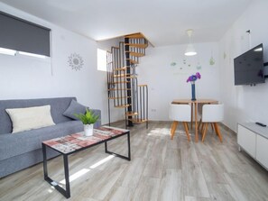 En moderne stue med en grå sofa, et sofabord med magasiner, en lille plante, en træspiral-trappe, et spiseområde med et hvidt bord og stole, samt et TV monteret på væggen.