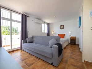 En moderne stue med en grå sofa, et lille bord og en seng med en orange pude. Der er et vindue med gardiner, en fastgjort klimaanlæg og en billedramme.