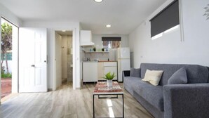 A compact living space with a grey sofa, a coffee table, a kitchen area, and a door leading to a balcony.