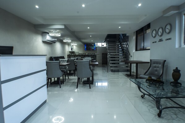 A modern hotel lobby with a reception desk, seating area, staircase, and a wall with clocks.