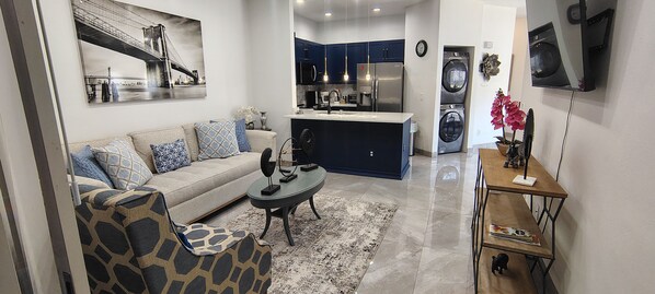 A modern living room with a sofa, coffee table, and patterned armchair. A kitchen with dark cabinets and a refrigerator is visible in the background.