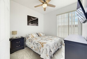 Une chambre à coucher comprenant un lit, une table de chevet, un ventilateur de plafond, une fenêtre avec des stores et un téléviseur fixé au mur.