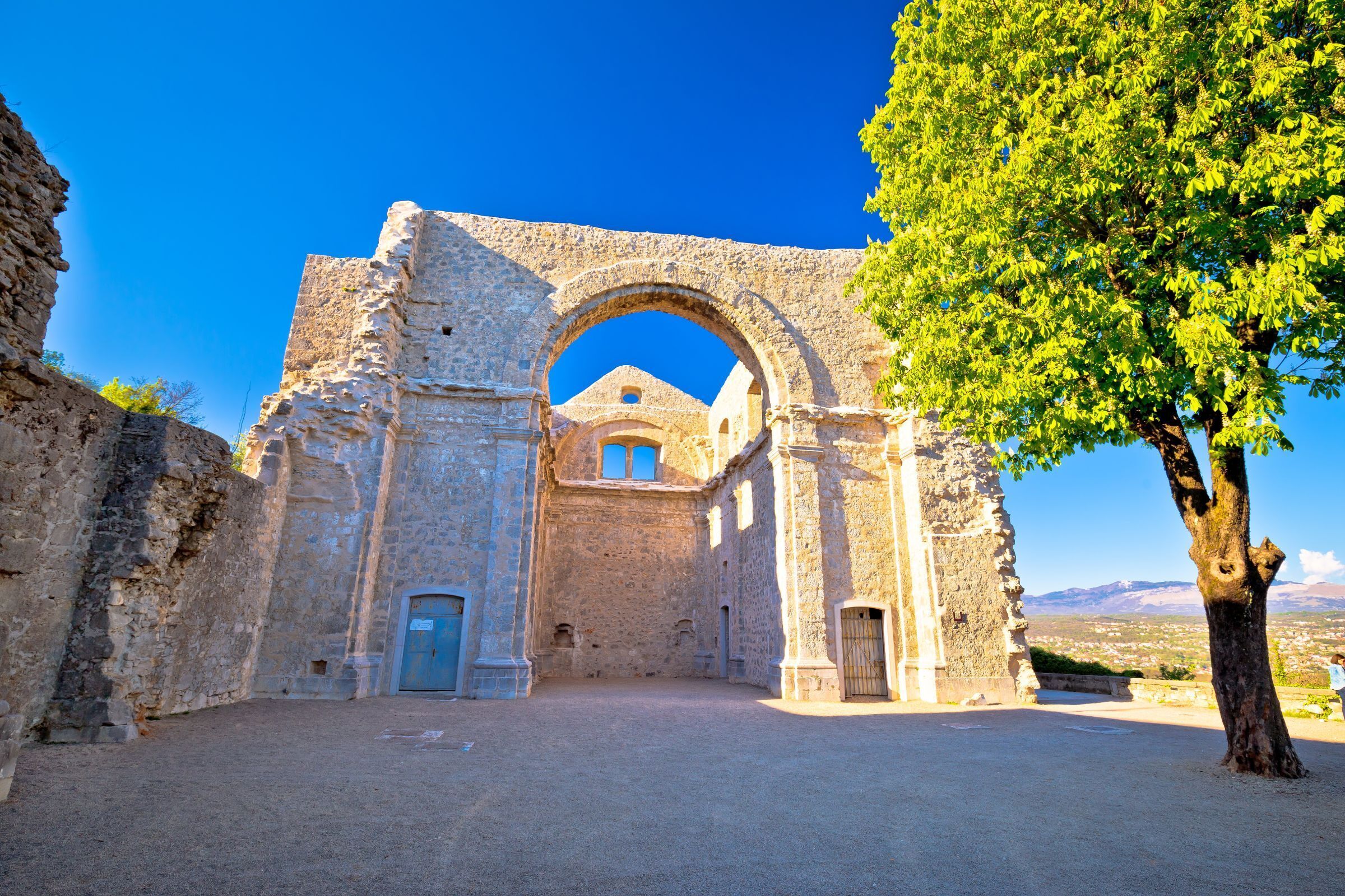 An ancient stone archway leading to a building with a blue door, surrounded by a clear sky and a tree.
