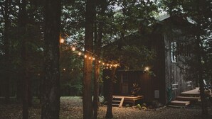 Una cabaña de madera rústica rodeada de árboles, con luces colgantes y un banco de madera.