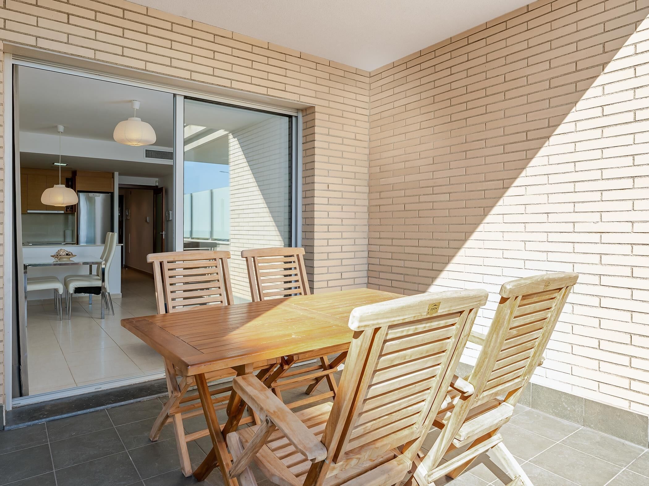 Un espacio moderno para comer al aire libre, con una mesa y sillas de madera, un ventanal de vidrio que da a una cocina y un muro de ladrillo.